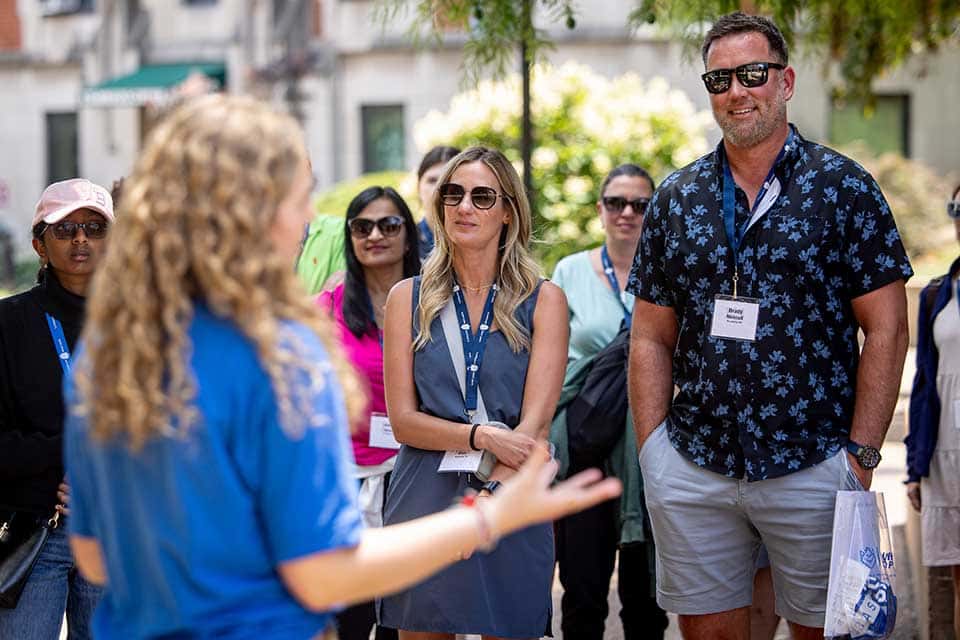 A group of parents stands in a circle as they listen to a female SLU Campus Ambassador speak during a campus tour. 