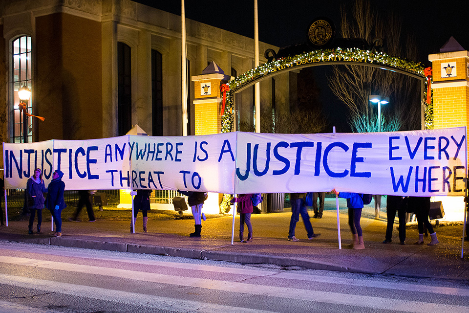D E I Office Students stand along a street on campus in front of an archway decorated by Christmas lights. They hold a long banner that reads "Injustice Anywhere is a Threat to Justice Everywhere"