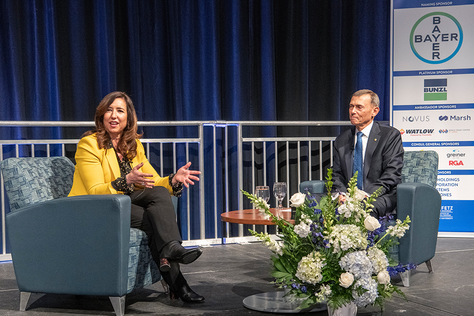 A woman sits on a stage while talking while a man in a suit looks on.