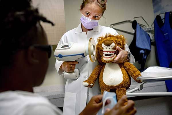 A female dentist demonstrates a dental tool on a stuffed animal with human teeth.