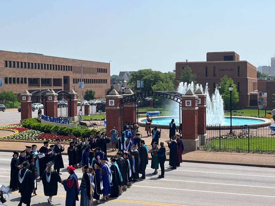 A group of faculty in academic regalia form a line and clap while graduates in caps and gowns cross the street. A bagpiper plays in the background.