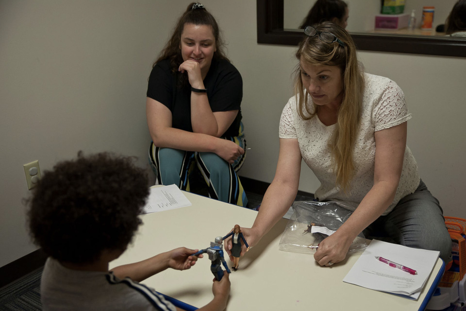 Autism Center A woman sits at a table across from a child, shown from the back. They each hold small toy figures toward each other. Another woman sits in the corner, smiling.