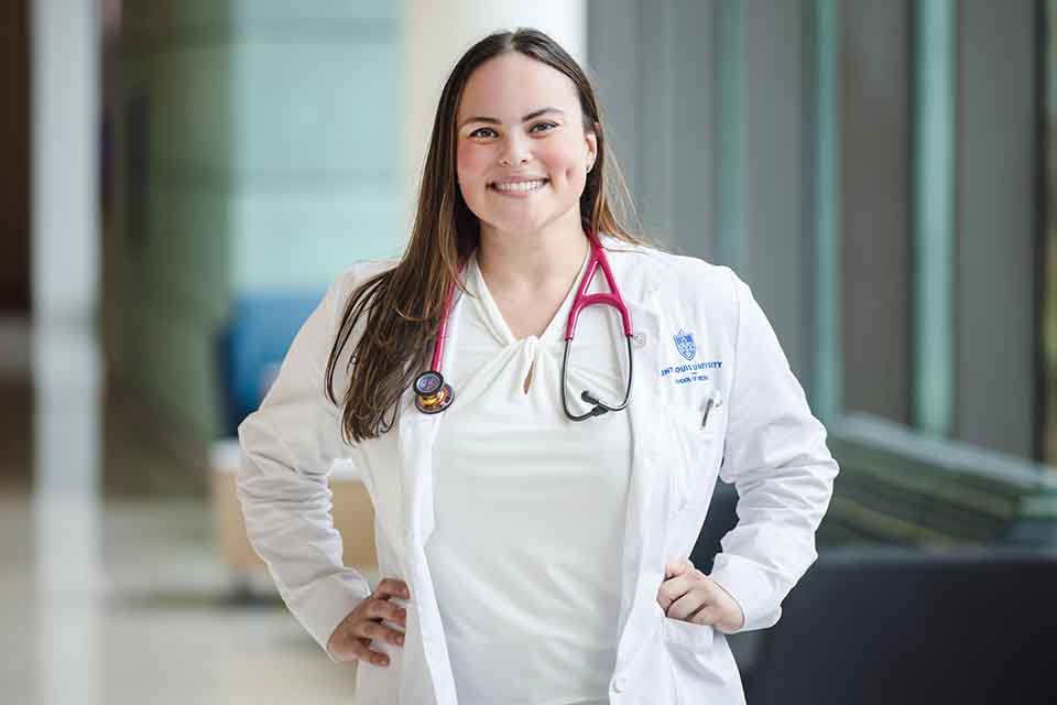 Caroline Wright Caroline Wright poses for a photo in the hallway of a medical building wearing a doctor's white coat.