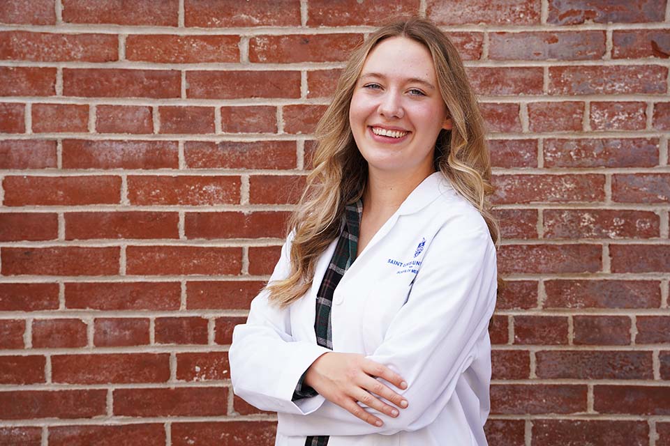 Caroline poses for a photo wearing her white lab coat against a brick wall.