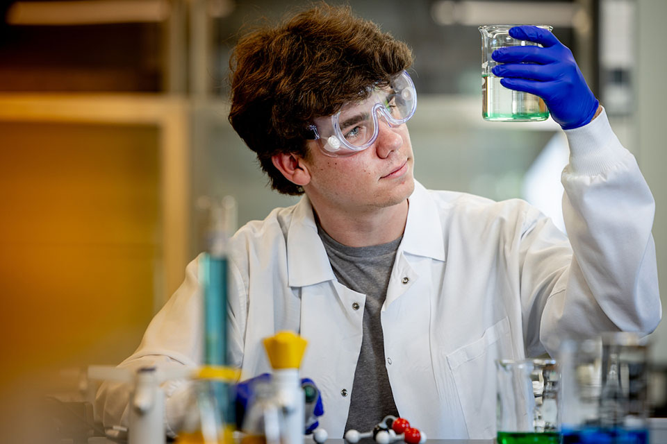 Eric Piening works in a lab, wearing a white lab coat and goggles.