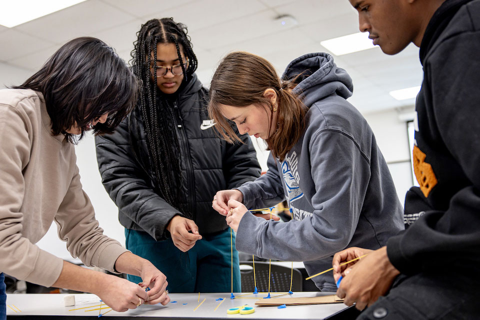 Engineering Four engineering students bend over a table working on a project involving wooden sticks.