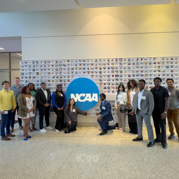 Students visit the NCAA and are posing in front of a NCAA wall with team logos.