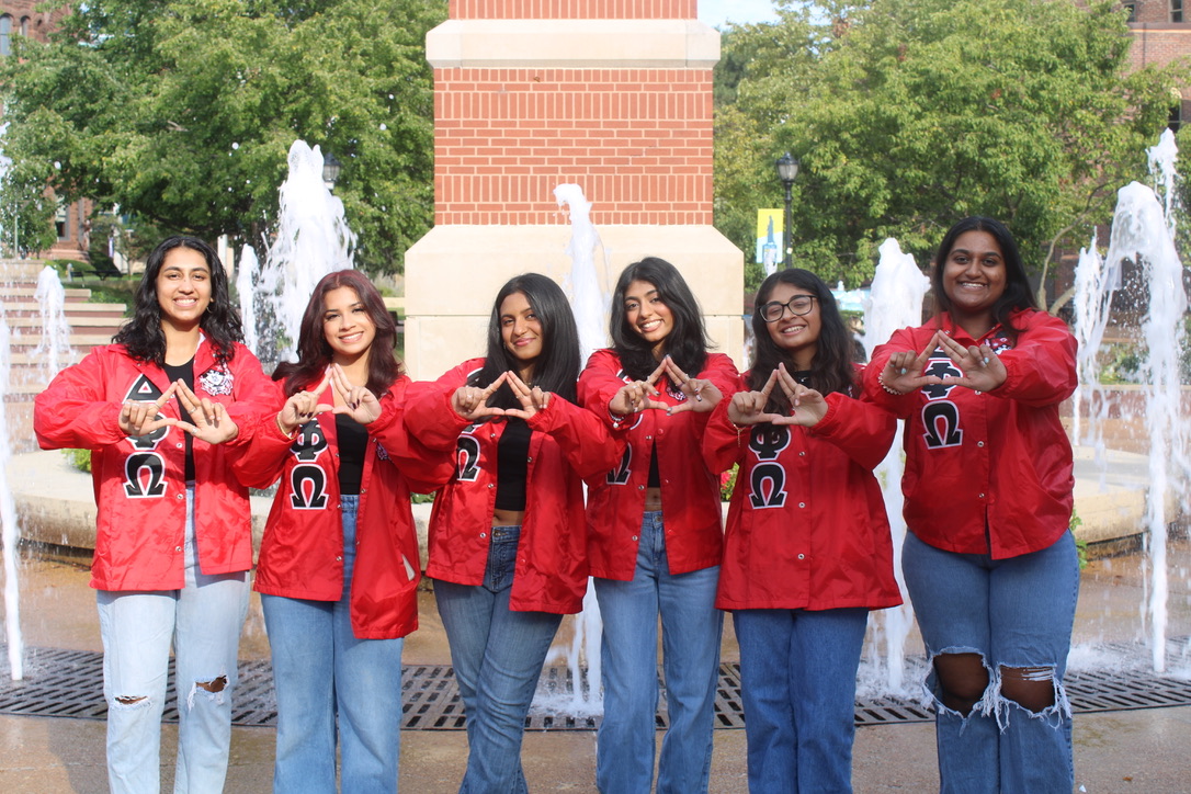 Sorority members wearing matching jackets with the greek letters for Delta Phi Omega make a delta symbol with their hands while standing in front of the SLU clock tower.