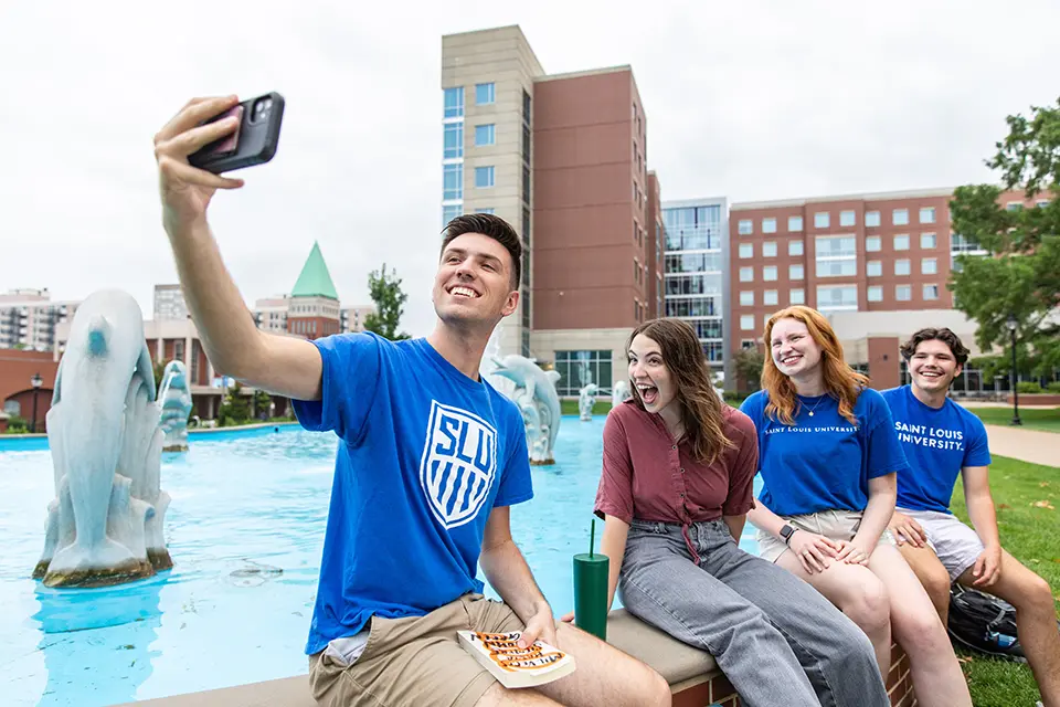 A student takes a selfie while his friends look at the camera, sitting in front of a fountain.