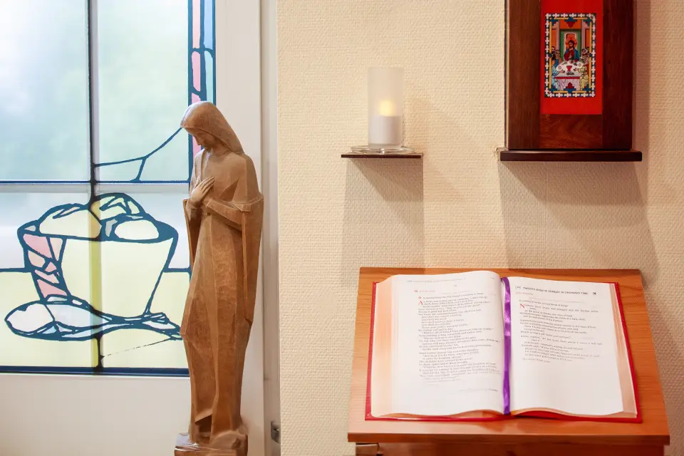 A wooden religious figure next to a candle and a bible in a room with a stained glass window.
