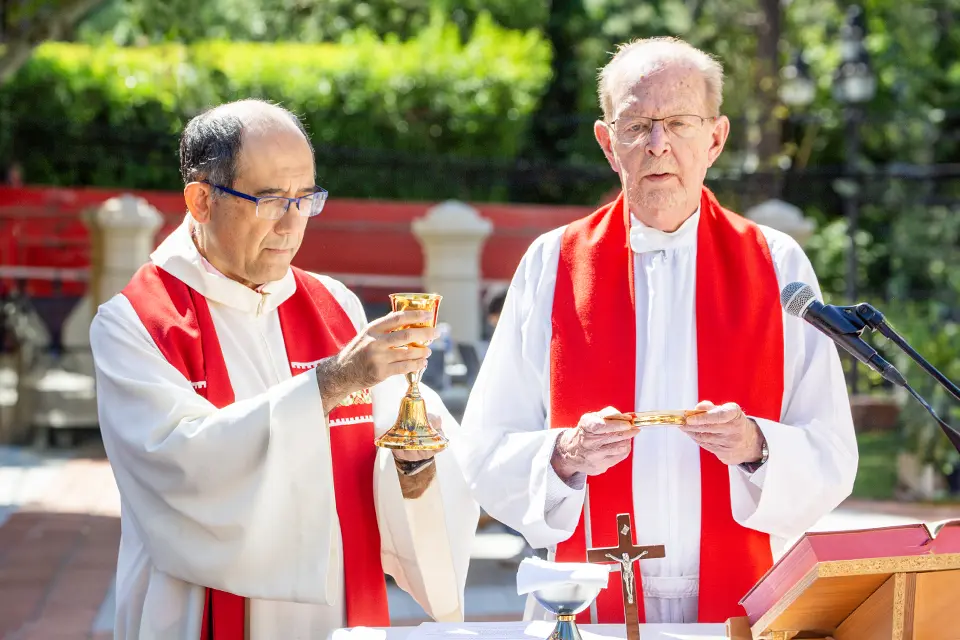 Two priests stand at an altar outside, while one holds a golden cup and another holds a golden plate.