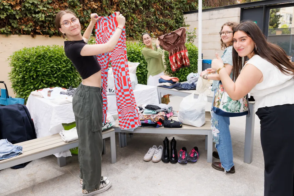 Students hold up articles of clothing and another student gives a thumbs up while standing around a table filled with clothing and shoes.