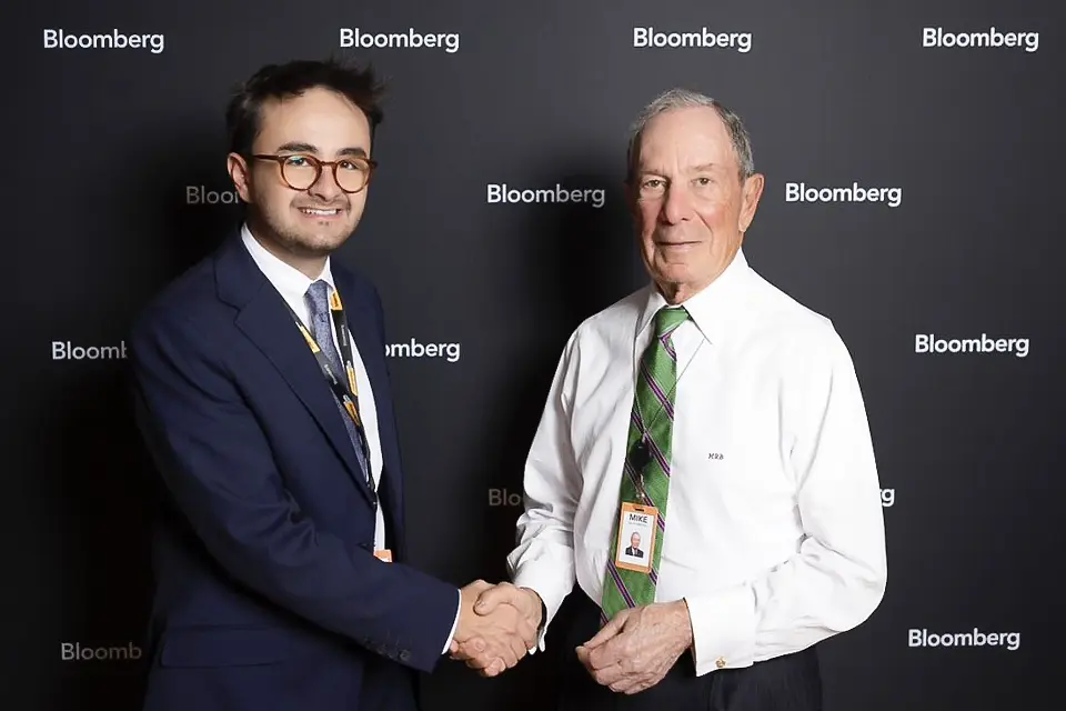 José Antonio Jijón shaking hands with Michael Bloomberg. An alumni shaking hands with a man in front of a photocall.