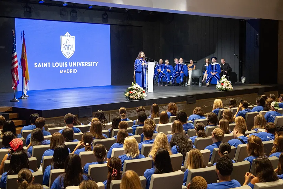 Co-interim director for academics Laurie Mazzuca, Ph.D., addresses incoming students in the Pablo VI Woman in academic regalia speaks to a crowd on stage in an auditorium full of students.