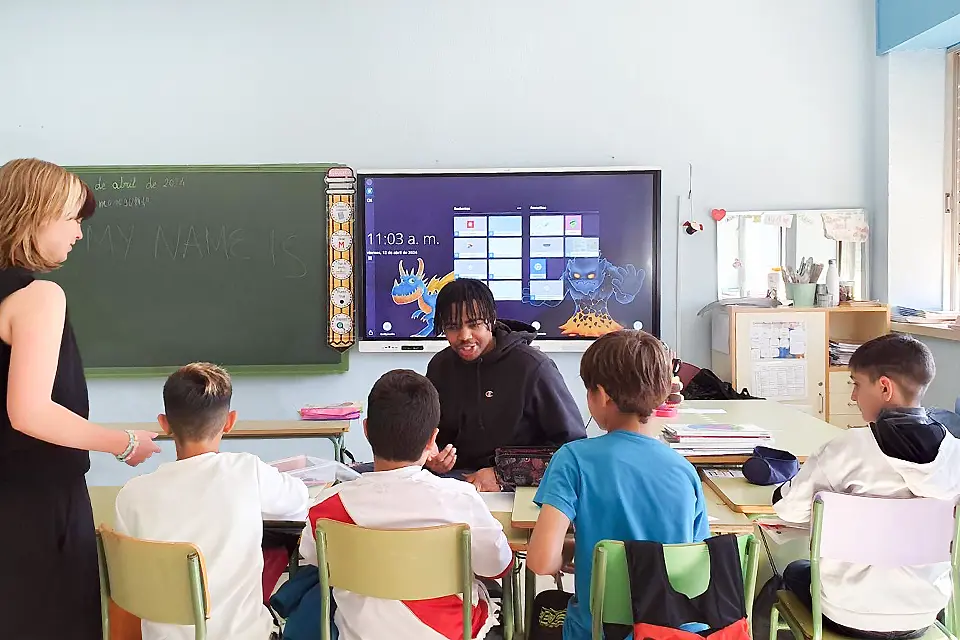 Student De’Von France sitting down with elementary schoolers during classroom visit. A SLU student sits talks to a group of four elementary students, seen from behind, sitting at desks in a classroom.