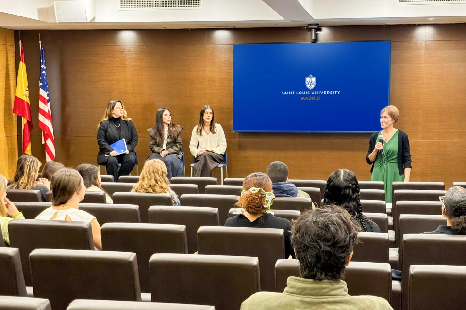 Career Services director Patrice Burns introduces alumni at a roundtable event on Dec. 11, 2025, in the San Ignacio Hall Auditorium. Three women sit on stage with Spain and U.S. flags to the left. There is a screen in front of the audience members and a woman dressed in a green dress holds a microphone and addresses the audience.