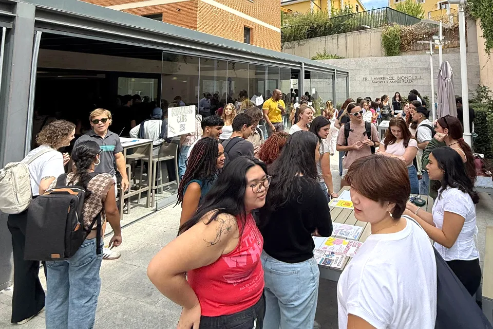 Many students standing around tables in an outdoor patio with a brick wall in the background and doors into a covered patio space.
