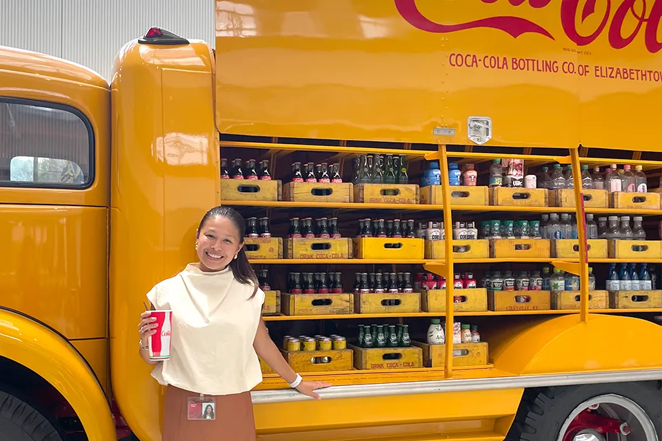 A young girl standing in front of a yellow truck holding a glass of soda in her hand.