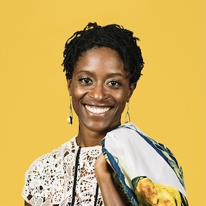 A shoulder-length photograph of an adult woman with tightly braided black hair wearing dangling earrings and a floral-lace pattern top, holding a floral-print wrap draped across her left shoulder. She stands in front of a canary yellow background and smiles warmly.
