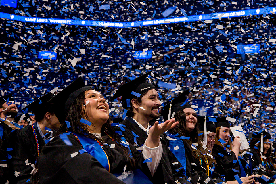 Graduates sit in chairs in the bowl of Chaifetz Arena. Confetti falls around them.