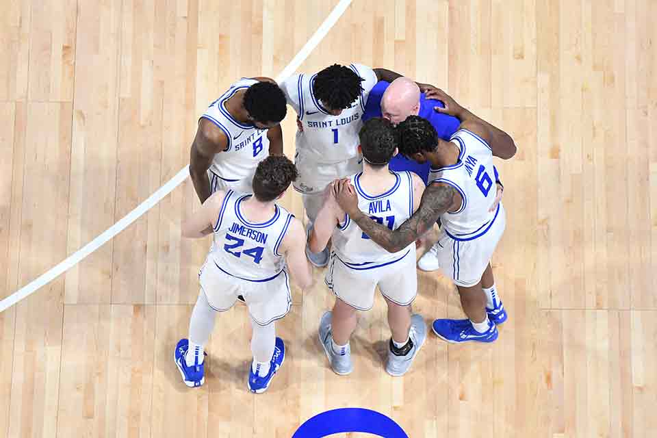 SLU Men's Basketball Members of the men's basketball team stand in a huddle in the middle of the court during a game.