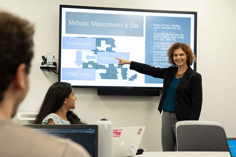 Geospatial Health A woman points to a TV screen that is displaying data from a recent study. Two students look on.