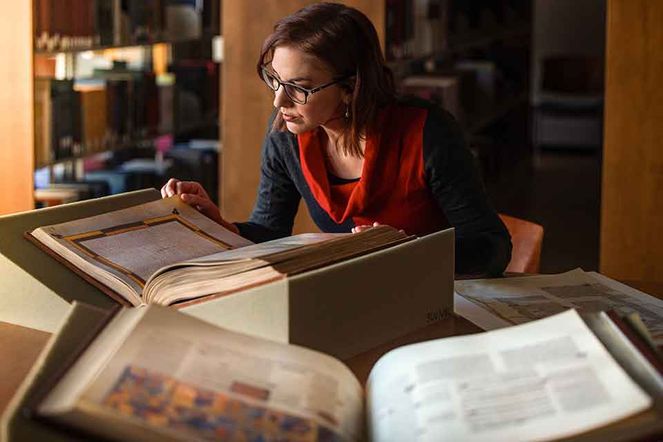 Humanities Unique Resources A woman leans over three illuminated manuscripts that are spread across a table top.
