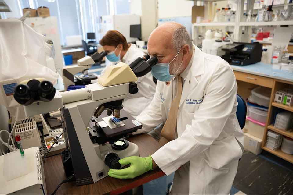 Two scientists wearing face masks as they examine samples through microscopes.