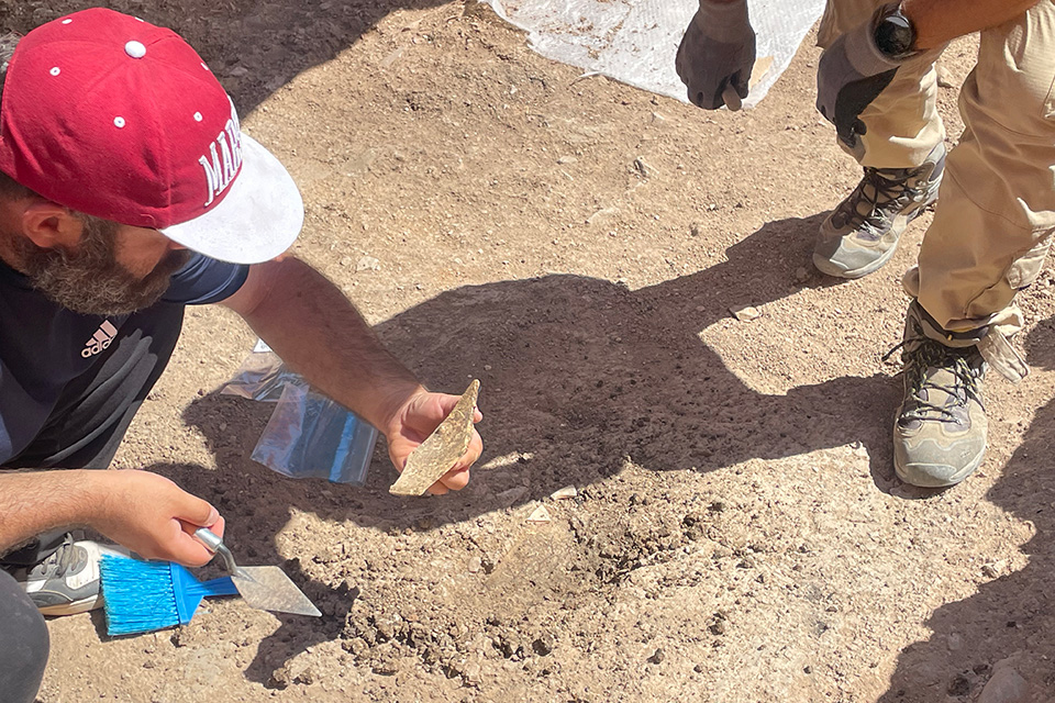 A team member holds a piece of the structure present at the archeological site. A team member holds a piece of brick or stone in one hand and an archeological implement in the other. The dusty, booted feet of a colleague are seen on the side of the image.
