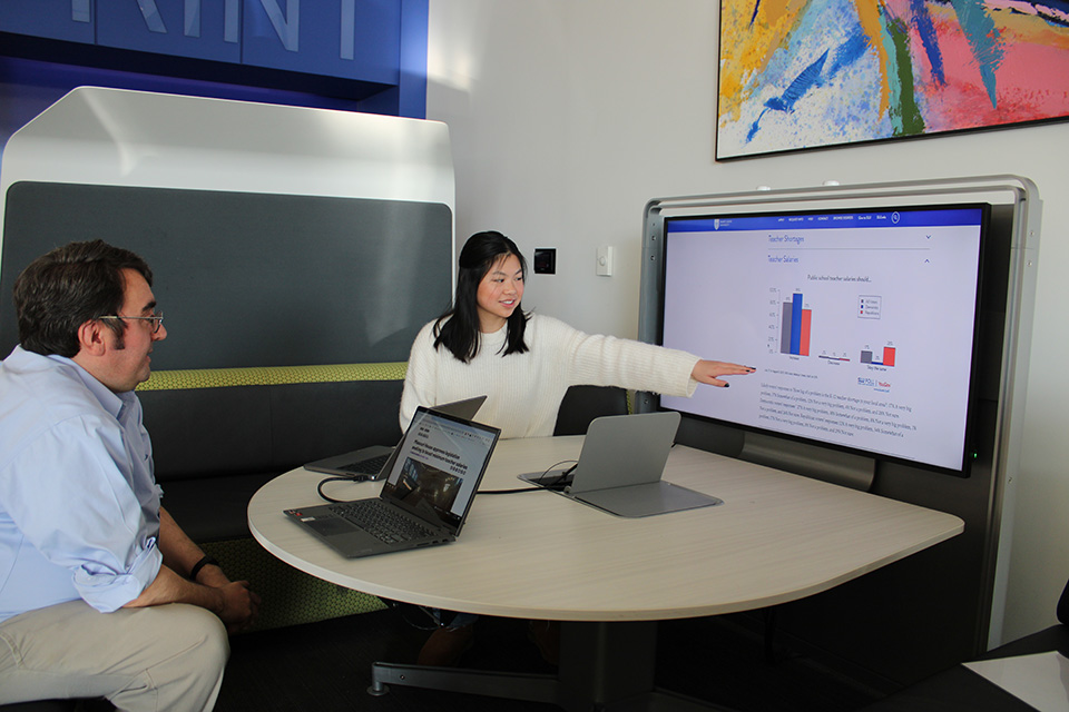 Klasen gestures toward a screen with bar graphs while Rogers looks on.