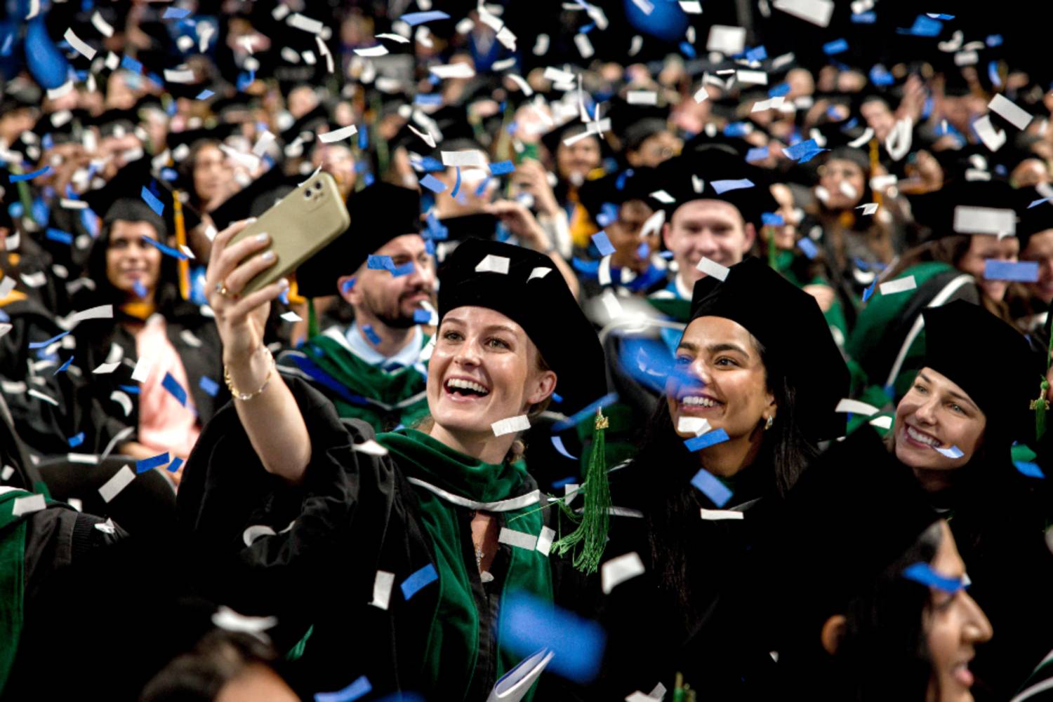 A student in graduation gear takes a selfie while confetti falls during the commencement ceremony.