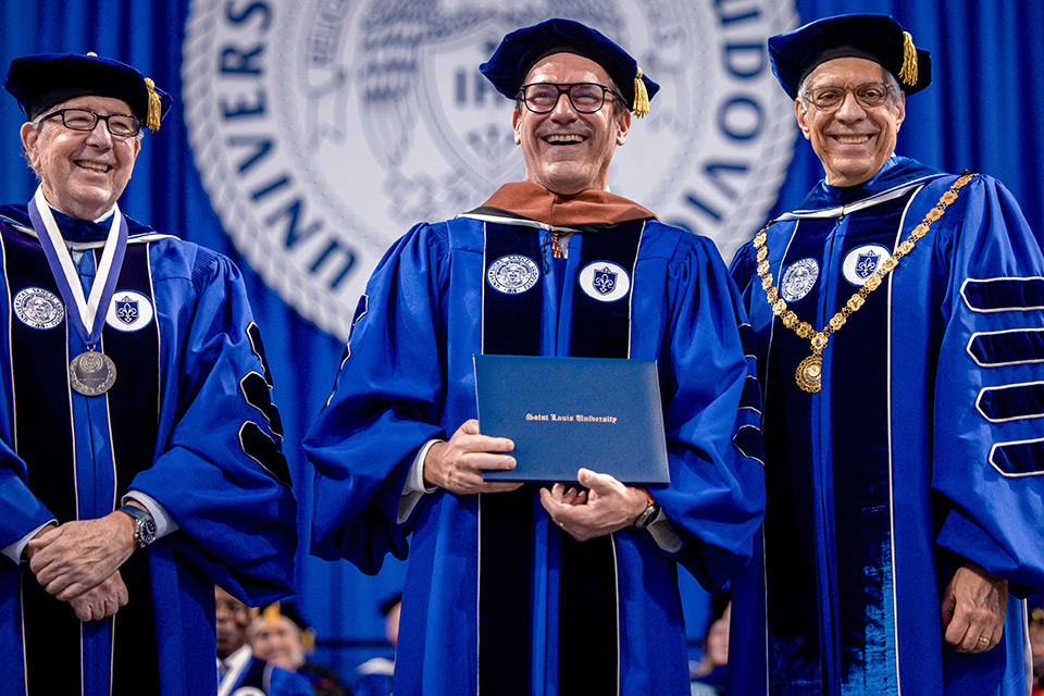 Jon Hamm holds a SLU diploma at the 2024 commencement ceremony with Joe Conran (left) and Pestello Actor Jon Hamm smiles as he holds a SLU diploma during the 2024 commencement ceremony. He is flanked by Joe Conran (left) and Fred Pestello (right). All men wear royal blue academic regalia.