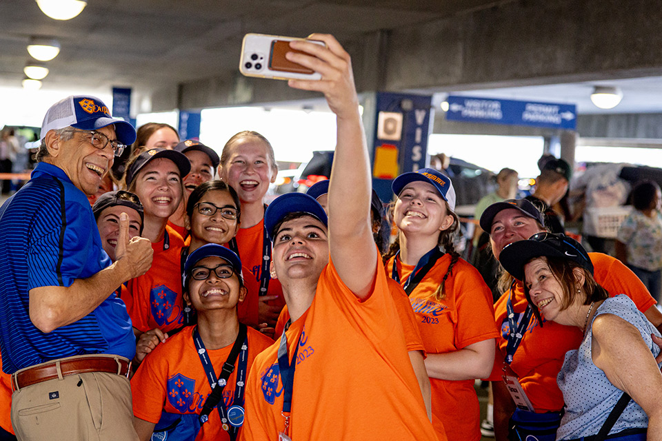 Pestello poses for a selfie with Oriflamme leaders during Fall Welcome. Pestello in a polo shirt and khakis poses for a selfie with numerous orange-shirted Oriflamme members in the Laclede Garage during Fall Welcome in 2023.